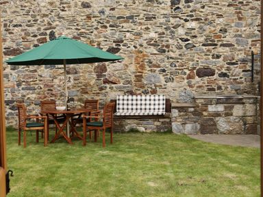 An outdoor seating area with wooden table and chairs under a green umbrella and a stone bench with cushion at Little Orchard in Ashburton Down near Ashburton