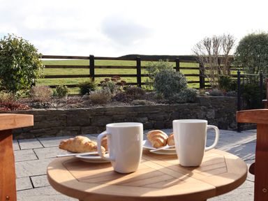 A wooden table with two white mugs and plates with croissants on a patio with garden and wooden fence in the background at Little Orchard in Ashburton Down near Ashburton