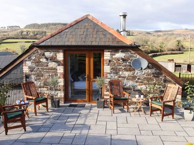 A stone house with wooden doors and four wooden chairs with cushions on a patio at Little Orchard in Ashburton Down near Ashburton