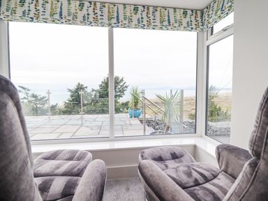 A sitting area with two gray recliner chairs facing large windows overlooking a patio with potted plants and trees at Aelfor in Harlech