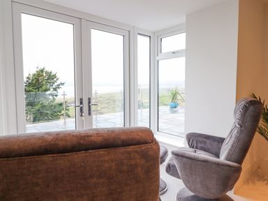 A sitting area with a brown sofa and gray chair facing glass doors and windows at Aelfor in Harlech