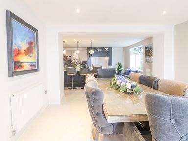 A dining area with a wooden table and upholstered chairs leading to a kitchen with bar stools and hanging lights at Aelfor in Harlech