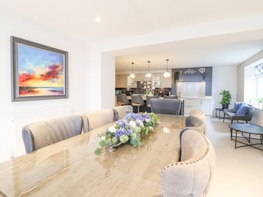 A dining area with upholstered chairs around a wooden table with a floral centerpiece and a kitchen in the background at Aelfor in Harlech