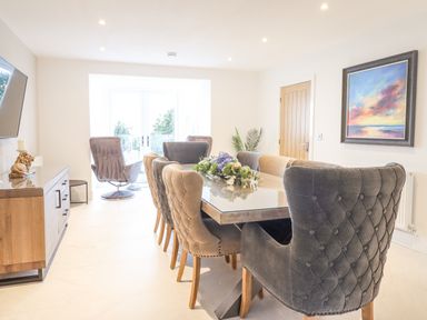 A dining room with a long table surrounded by upholstered chairs a sideboard a wall painting and glass doors at Aelfor in Harlech