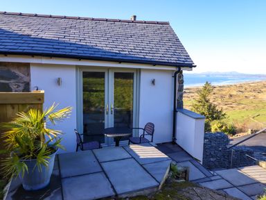 A small outdoor patio with a round table and two chairs outside a building with double glass doors and a potted palm plant overlooking a coastal landscape at Aelfor in Harlech