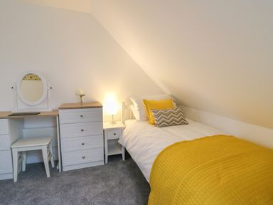 A bedroom with a single bed with cushions and yellow bedding a bedside table with a lamp a chest of drawers and a dressing table with a stool at 2 Penporth in Rhayader