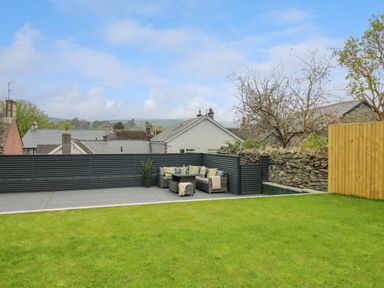 A backyard with a green lawn a grey patio a corner seating area with cushions and a stone wall at 2 Penporth in Rhayader