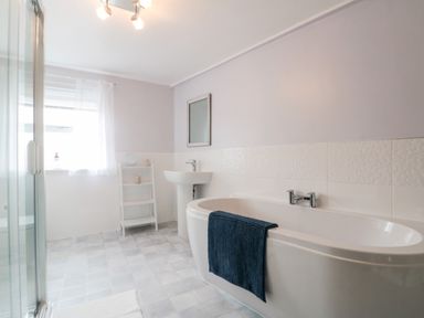 A bathroom with a bathtub with a towel a sink a mirror a glass shower a window with curtains and a shelf at Bluebell Cottage in Flamborough