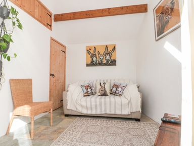 A small room with a sofa covered by a blanket and animal print cushions a wicker chair a wooden door a hanging plant and animal paintings at Bluebell Cottage in Flamborough