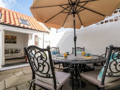 An outdoor patio with a metal table and chairs under a large umbrella and an open door leading to a room with a sofa and animal artwork at Bluebell Cottage in Flamborough