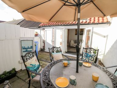 An outdoor patio with a round table under a large umbrella four chairs with cushions and plates and mugs on the table at Bluebell Cottage in Flamborough