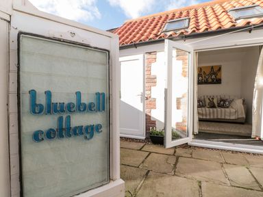 An entrance to a cottage with a sign reading bluebell cottage and an open door showing a room with a sofa and animal themed cushions at Bluebell Cottage in Flamborough