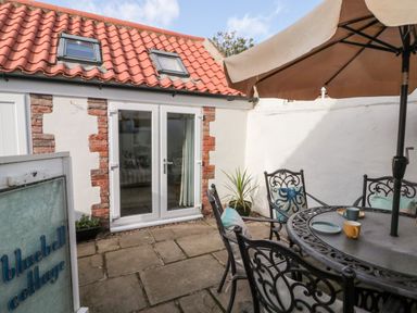 An outdoor patio area with a round metal table and four chairs with cushions under a large umbrella at Bluebell Cottage in Flamborough