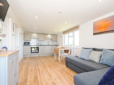 A kitchen and living room area with wooden floors a grey sofa with pillows a dining table and chairs and kitchen cabinets at Lundy View Chalet in Widemouth Bay