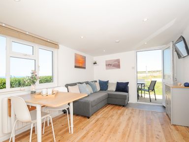 A living room with a dining table and chairs a gray corner sofa and an open door leading outside at Lundy View Chalet in Widemouth Bay