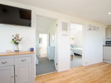 A room with wooden floor a gray cabinet with flowers on it two doorways leading to carpeted bedrooms a wall-mounted TV and kitchen cabinets at Lundy View Chalet in Widemouth Bay