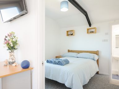 A bedroom with a bed with blue bedding and rolled towels with a vase of flowers on a wooden surface and a wall-mounted TV at Lundy View Chalet in Widemouth Bay