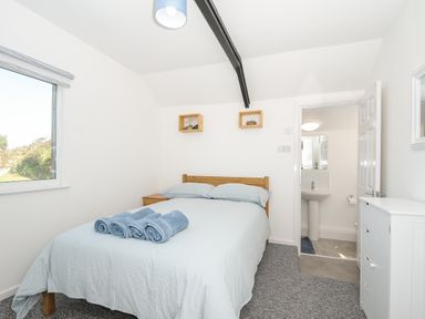 A bedroom with a double bed with blue linens towels on top bedside table window and doorway leading to a bathroom at Lundy View Chalet in Widemouth Bay