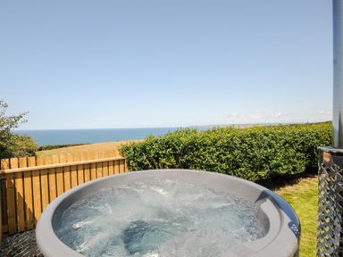An outdoor hot tub with water bubbles a wooden fence bushes and the sea in the background at Lundy View Chalet in Widemouth Bay