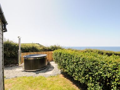An outdoor hot tub on gravel near a wooden fence with a hedge and sea view at Lundy View Chalet in Widemouth Bay