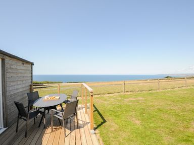 An outdoor deck with a table and four chairs overlooking a grassy field and the sea at Lundy View Chalet in Widemouth Bay