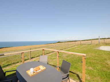 A patio table with chairs and croissants on a tray overlooking a fenced grassy area and the sea at Lundy View Chalet in Widemouth Bay