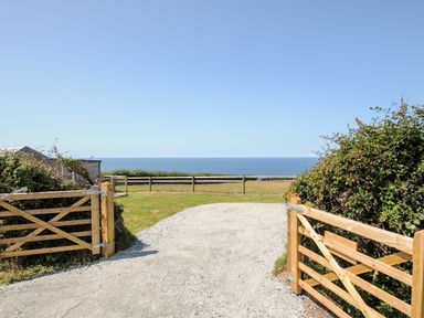 A gravel driveway with wooden gates opening to a grassy area and a view of the sea and clear sky at Lundy View Chalet in Widemouth Bay
