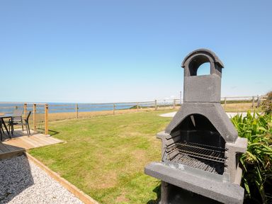 An outdoor area with a stone barbecue grill on the grass and a deck with chairs overlooking the ocean at Lundy View Chalet in Widemouth Bay