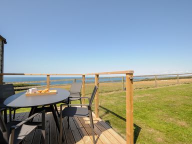 An outdoor deck with a table and chairs overlooking a field and the sea at Lundy View Chalet in Widemouth Bay