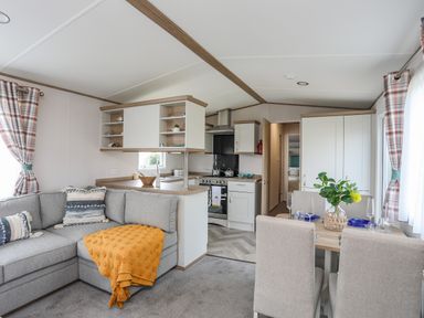 A living room with a gray sectional sofa a dining table with four chairs and a kitchen area at Moondunes - Pwllheli in Chwilog