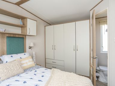 A bedroom with a bed featuring sea-themed bedding and pillows a wardrobe with drawers an open door showing a bathroom with a toilet at Moondunes - Pwllheli in Chwilog