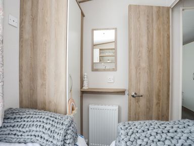 A bedroom corner with two wooden doors a small wooden shelf with a decorative lighthouse a mirror above a radiator and thick knitted gray blankets on beds at Moondunes - Pwllheli in Chwilog