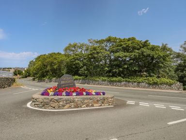 A stone roundabout with flowers and a sign reading Ocean Heights Caravan Park near a road with trees and stone walls in the background at Moondunes - Pwllheli in Chwilog