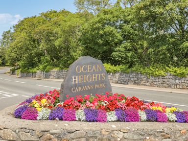 A stone sign reading Ocean Heights Caravan Park surrounded by colorful flowers beside a road with trees and a stone wall in the background