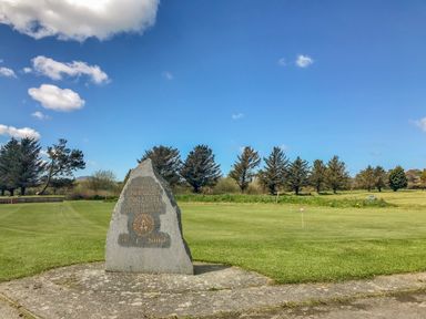 A stone memorial on a grassy golf course with trees and a blue sky in Pwllheli