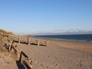 A sandy beach with wooden railings and a walkway next to the sea and hills in the distance at Moondunes - Pwllheli in Chwilog