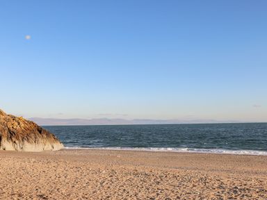 A sandy beach with a rocky cliff on the left and the sea with waves under a clear blue sky at Moondunes - Pwllheli in Chwilog