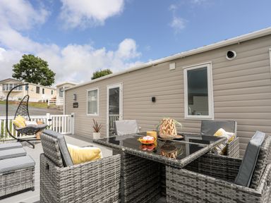 An outdoor patio area with wicker chairs around a glass table and a hanging wicker chair attached to a beige mobile home at Moondunes - Pwllheli in Chwilog