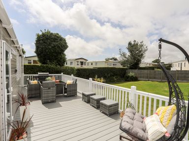 An outdoor deck with wicker furniture a hanging chair and a barbecue grill overlooking a grassy fenced garden at Moondunes - Pwllheli in Chwilog