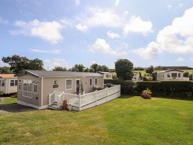 A caravan with a white fenced deck on a grassy plot under a partly cloudy sky at Moondunes - Pwllheli in Chwilog