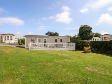 A mobile home with a white deck and lawn surrounded by trees at Moondunes - Pwllheli in Chwilog