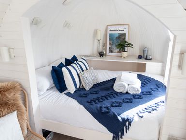 A bed with blue and white pillows and a blue blanket with rolled white towels near a shelf with a plant and picture at Andrus - Mayflower Meadow in Brandesburton