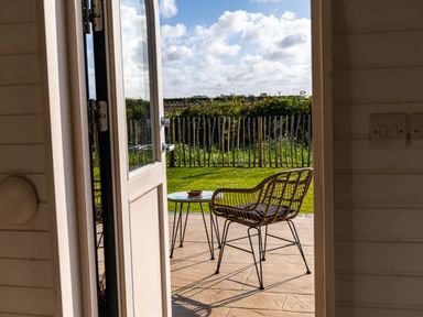An open door leading to a patio with a metal table and a woven chair overlooking a garden with a wooden fence at Indrek - Mayflower Meadow in Brandesburton