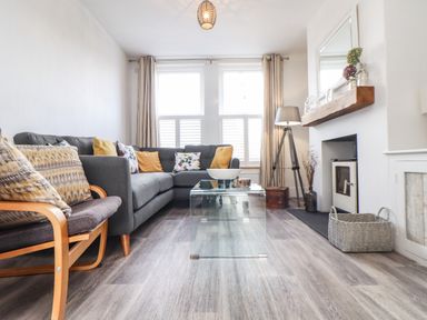 A living room with a gray sectional sofa with cushions a glass coffee table a wooden chair with cushions a floor lamp a fireplace and a basket at Chy An Mor in Mevagissey