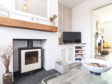 A living room with a wood burning stove a TV on a white cabinet and a glass table with a decorative bowl at Chy An Mor in Mevagissey