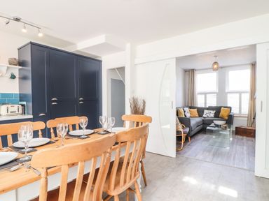 A dining area with wooden chairs and a table set with plates and glasses next to a living room with a gray sofa and glass coffee table at Chy An Mor in Mevagissey