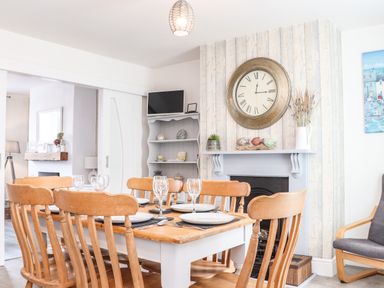 A dining area with a wooden table and six chairs set with plates and glasses near a fireplace and a large wall clock at Chy An Mor in Mevagissey