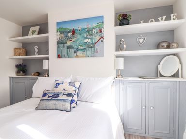 A bedroom with a bed with fish patterned pillows two grey cupboards with shelves above with lamps and decorations at Chy An Mor in Mevagissey