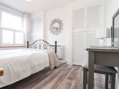 A bedroom with a bed metal headboard white bedding a window with shutters built in cupboards a dressing table with a lamp and mirror at Chy An Mor in Mevagissey
