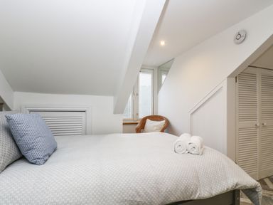 A bedroom with a bed with pillows and rolled towels a wicker chair near windows and closets with louvered doors at Chy An Mor in Mevagissey
